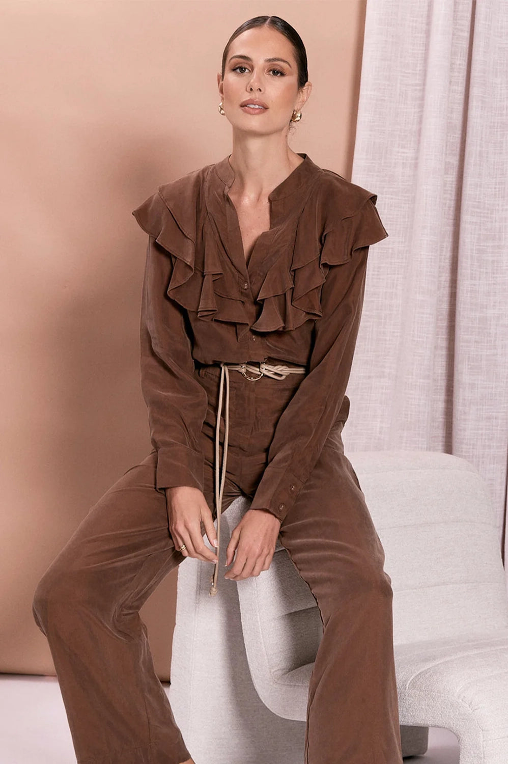 Woman wearing a brown ruffled top and pants sitting on a white chair.